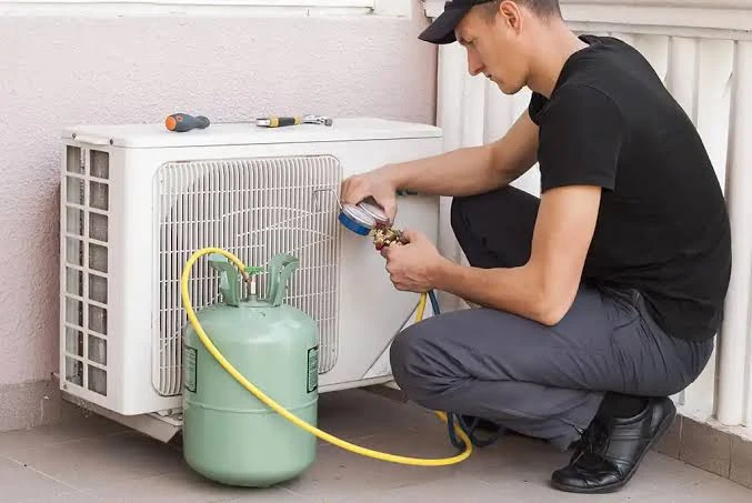 Technician servicing a wall-side air conditioning unit with a refrigerant cylinder and gauges